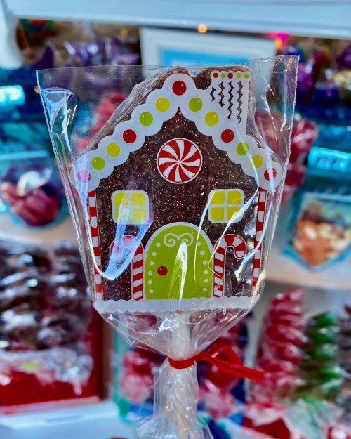 A colorful gingerbread house lollipop wrapped in clear plastic, with festive decorations and a red ribbon.
