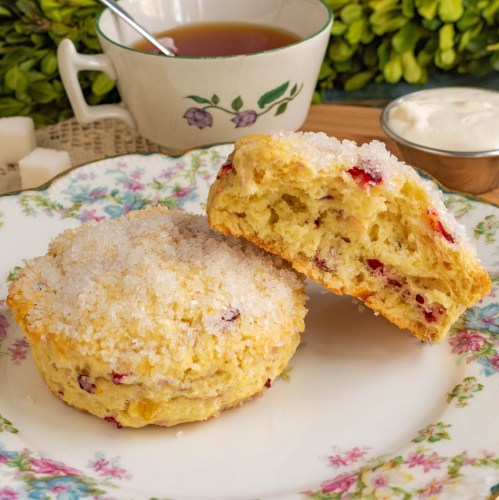 A close-up of a sweet scone topped with sugar, with a cup of tea and cream in the background on a floral plate.