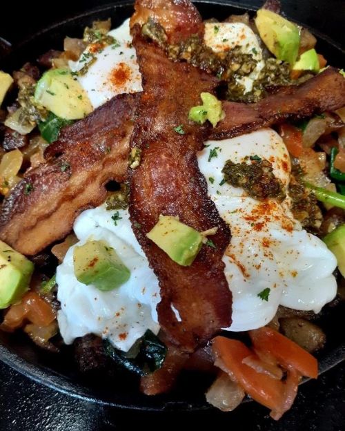 A skillet filled with poached eggs, crispy bacon, avocado, and colorful vegetables, garnished with herbs and spices.