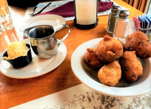 A bowl of golden fried fritters next to a cup of coffee and a small dish of butter on a wooden table.