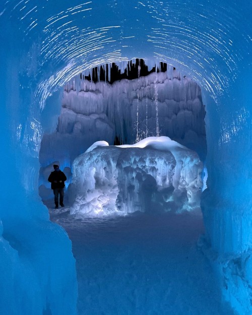 A person stands in a stunning ice cave, illuminated by blue light, with icy formations and stalactites above.