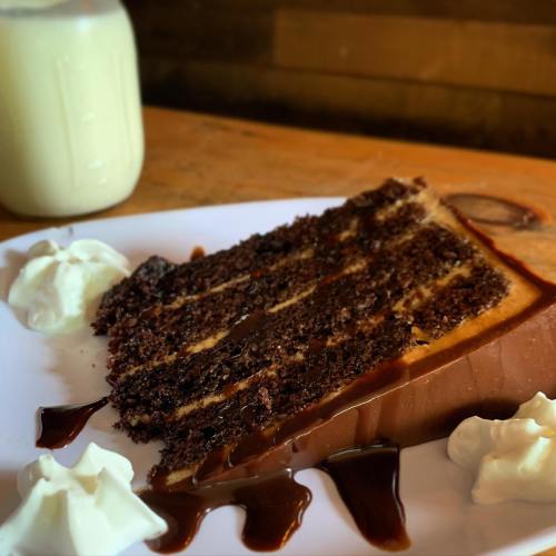 A slice of chocolate cake drizzled with chocolate sauce, accompanied by whipped cream and a glass of milk.