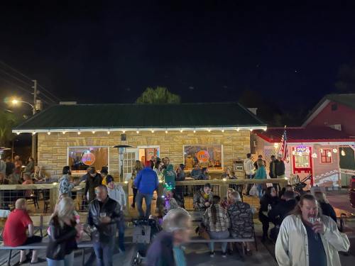 A lively outdoor scene at night with a crowd gathered in front of a stone building, enjoying drinks and conversation.