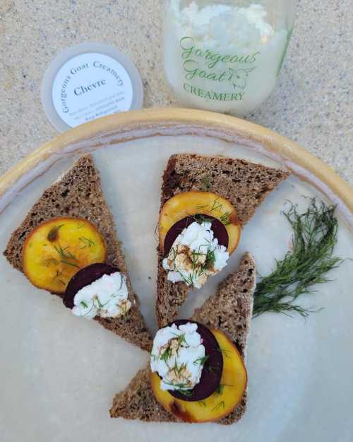 Sliced rye bread topped with goat cheese, beets, and peach, served on a plate with a jar of goat cheese nearby.