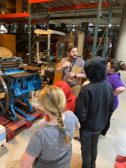 A guide explains machinery to a small group of people in a workshop setting, surrounded by various equipment.