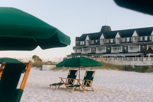 Two green beach chairs under umbrellas on a sandy beach, with a hotel in the background.
