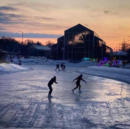 Skaters glide on an outdoor ice rink at sunset, with colorful lights and a greenhouse in the background.