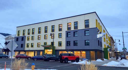 Modern multi-story building with colorful windows, parked cars in front, and a snowy landscape in the background.
