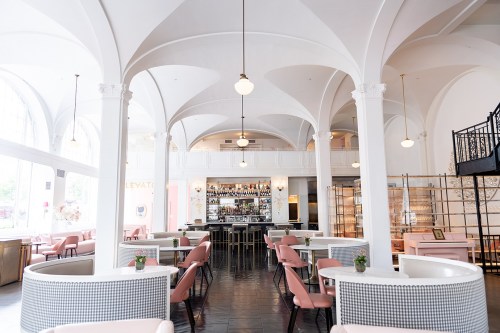 A stylish restaurant interior featuring arches, pink seating, and a bar in the background, with large windows for natural light.