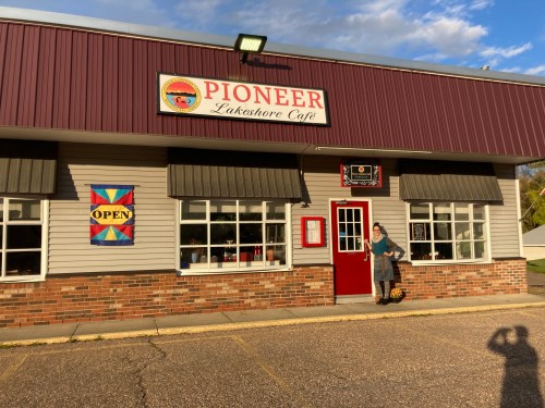 A woman stands outside the Pioneer Lakeshore Café, featuring a red door and colorful signs, under a blue sky.