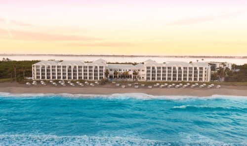 A beachfront hotel with white architecture, surrounded by palm trees and ocean waves, under a pastel sunset sky.