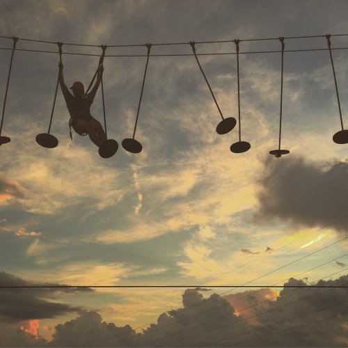 A person swings on a rope bridge against a colorful sunset sky with clouds.