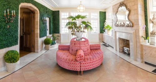 A stylish room with a pink patterned sofa, greenery on the walls, and elegant decor, featuring a fireplace and large windows.