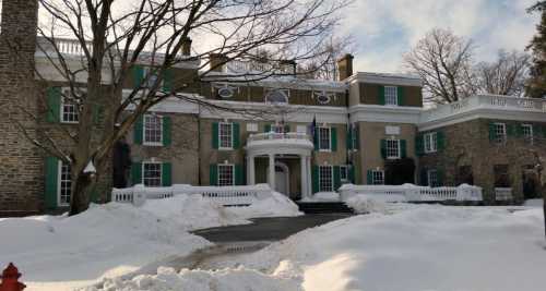 Historic building with green shutters, surrounded by snow, featuring a grand entrance and flags in winter.