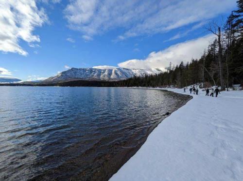 A snowy shoreline beside a lake, with mountains in the background and people walking along the water's edge.