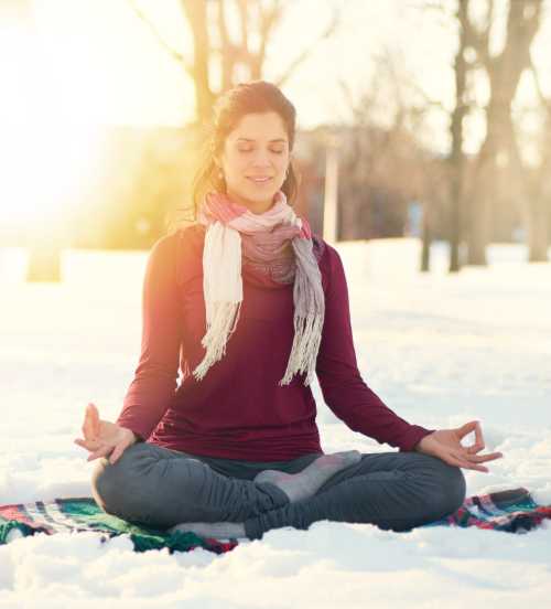 A woman meditating in the snow, sitting cross-legged on a blanket, with a serene expression and a scarf around her neck.