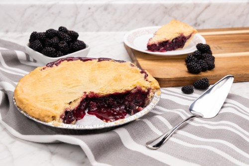 A blackberry pie with a slice removed, served with fresh blackberries on a gray striped tablecloth.