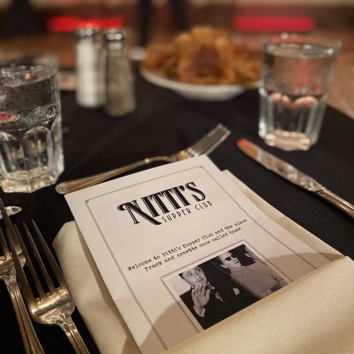 A table set with a menu for Kitt's Supper Club, glasses of water, and a plate of food in the background.