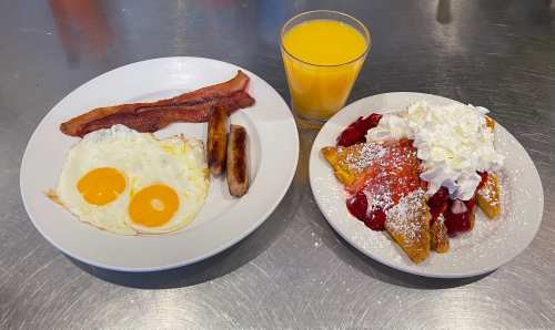 A plate with eggs, bacon, and sausage beside a plate of French toast topped with strawberries, whipped cream, and a glass of orange juice.