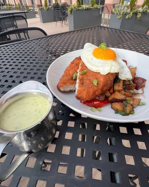 A plate of crispy fried fish topped with a sunny-side-up egg, served with potatoes and a small cup of green sauce.