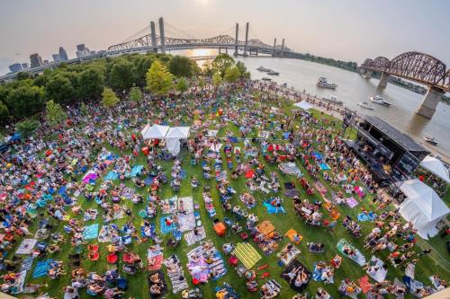 Aerial view of a large crowd enjoying a picnic on the grass by a river, with bridges and a stage in the background.
