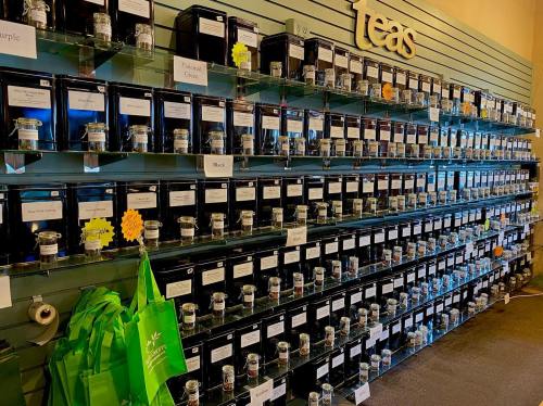 A wall display of various tea jars, labeled with names, organized on shelves in a tea shop. Green bags are visible below.