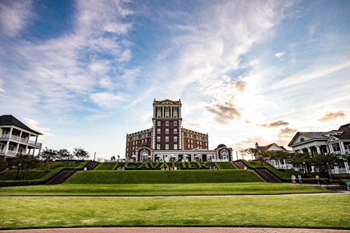 A grand hotel with a tower stands against a colorful sky, surrounded by green lawns and walking paths.