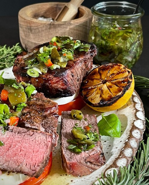 Grilled steak slices topped with green sauce, served with charred lemon and fresh herbs on a decorative plate.