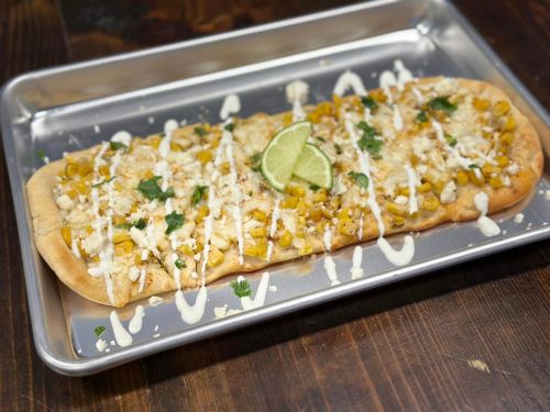 A flatbread topped with corn, cheese, cilantro, and lime, drizzled with sauce, served on a metal tray.