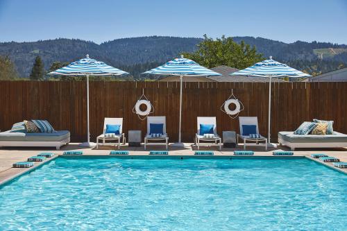 A sunny pool area with blue-striped umbrellas, lounge chairs, and a wooden fence, set against a mountain backdrop.