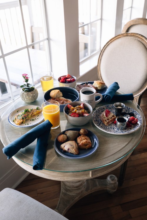 A beautifully arranged breakfast table with various dishes, including fruit, pastries, and drinks, by a window.