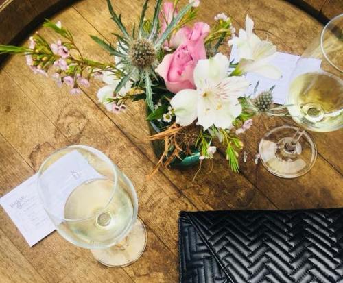 A wooden table with two glasses of white wine, a floral arrangement, and a black textured clutch purse.