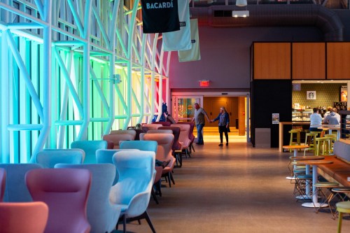 A modern interior with colorful lighting, featuring seating areas and a bar in the background. Two people walk hand in hand.