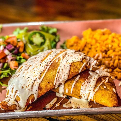 Two crispy tacos drizzled with sauce, served with rice, salad, and fresh toppings on a pink plate.