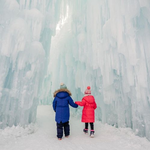 Two children in winter coats walk hand in hand through a stunning ice cave with towering icicles.