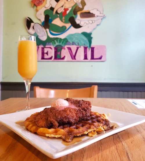 A plate of chicken and waffles with a mimosa in the background, featuring a colorful mural on the wall.
