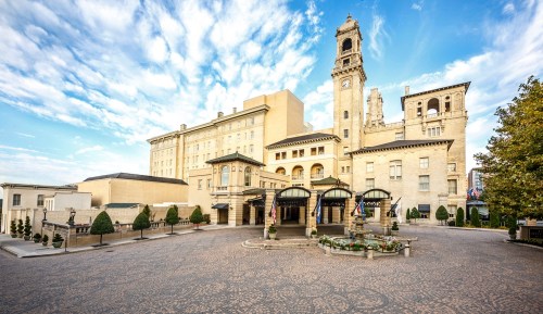 Historic building with a clock tower, surrounded by trees and a cobblestone entrance under a blue sky.
