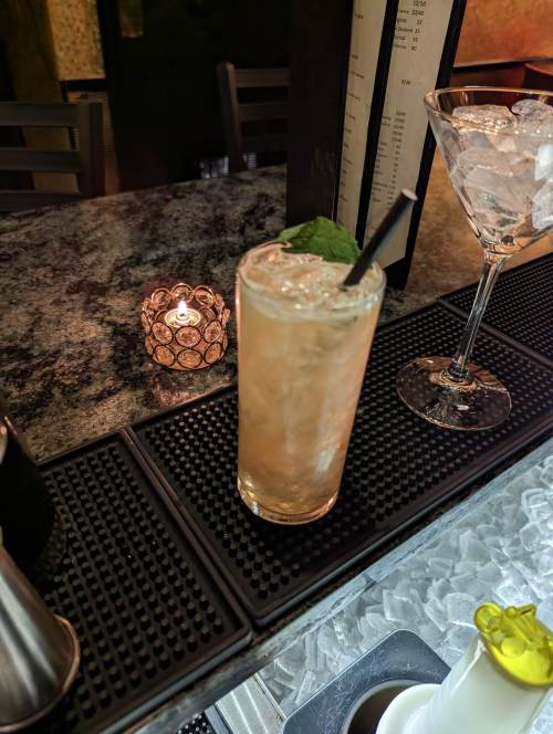 A refreshing cocktail with ice and mint sits on a bar counter next to a glass of ice and a lit candle.