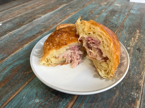 A flaky croissant cut in half, revealing shredded meat inside, on a decorative white plate against a rustic wooden table.
