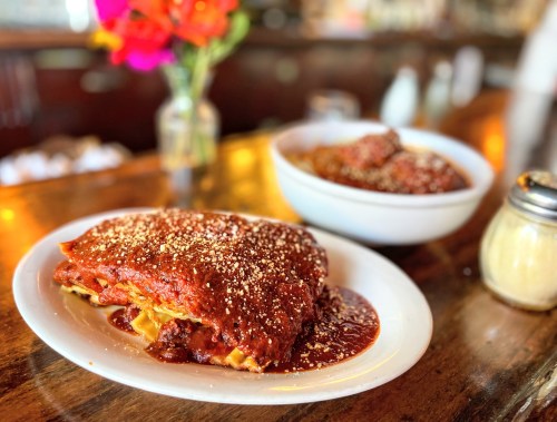 A plate of lasagna topped with marinara sauce and cheese, with a bowl of meatballs in the background and flowers on the table.