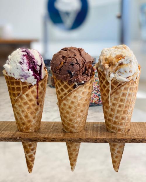 Three ice cream cones on a wooden holder: one with blueberry, one with chocolate, and one with caramel swirl.