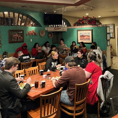 A lively restaurant scene with diverse groups of people dining at wooden tables, enjoying food and drinks.