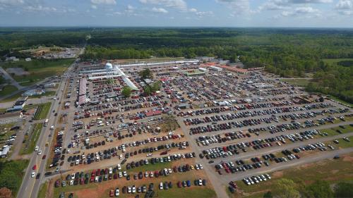 Aerial view of a large outdoor market with numerous cars parked and green trees in the background under a blue sky.
