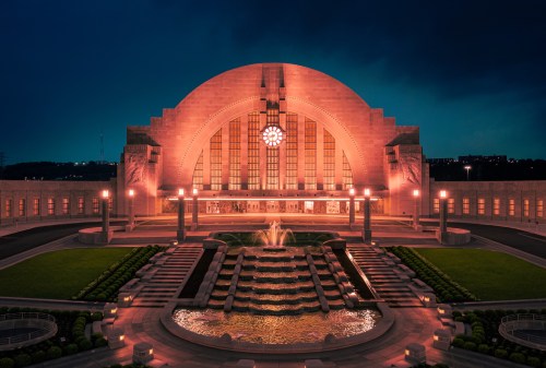 Historic building illuminated at dusk, featuring a large arched roof, decorative windows, and a central fountain.