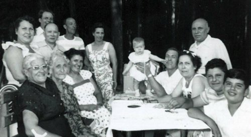 A black-and-white photo of a large family gathering around a table in a forested area, with adults and children smiling.
