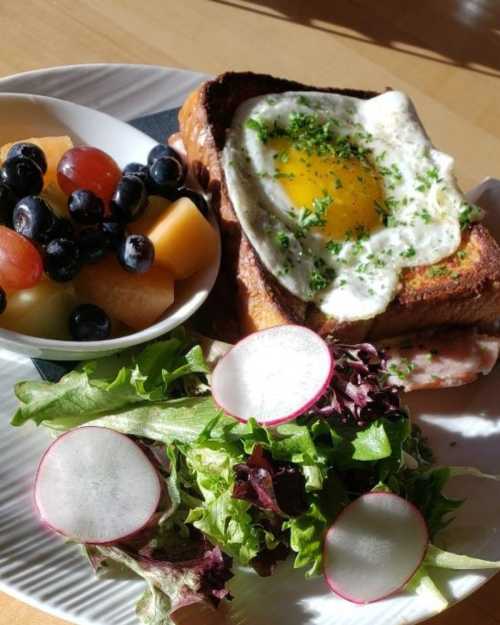 A plate with French toast topped with a fried egg, a side of mixed fruit, and a fresh salad with radishes.