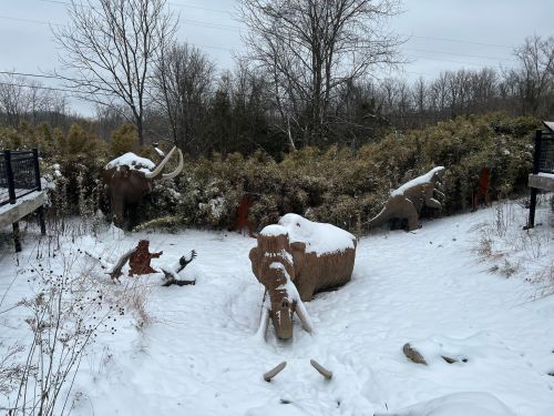 Snow-covered prehistoric animal sculptures in a winter landscape, surrounded by trees and shrubs.