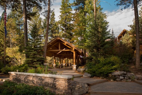 A rustic lodge entrance surrounded by tall trees and greenery, with a stone sign reading "The Cribs Resort."