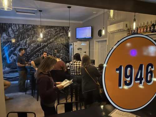 A cozy bar scene with patrons enjoying drinks, a large screen displaying content, and a view of the bar from outside.
