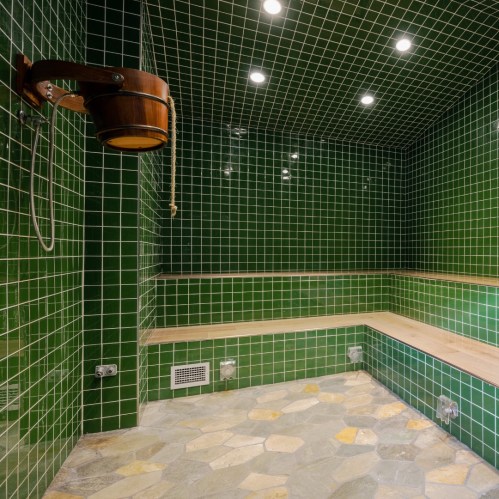 A modern sauna with green tiled walls, wooden benches, and bright overhead lights.